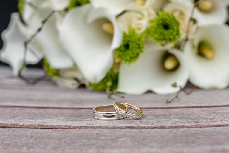 Close-up of wedding rings on wooden surface with white and green floral arrangement.